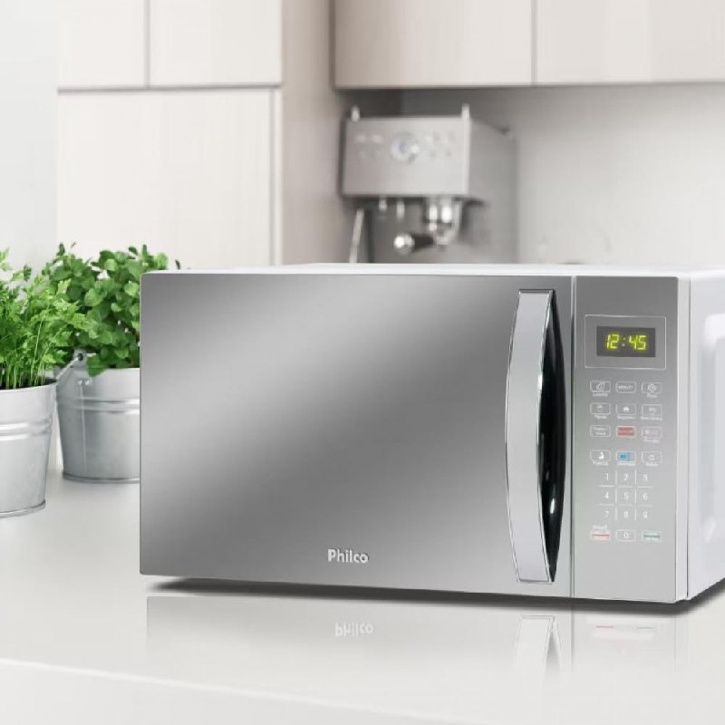 Micro-ondas Philco PMO38E de 33 litros na cor prata, com painel digital e porta espelhada, posicionado sobre uma bancada de cozinha ao lado de vasos com plantas.