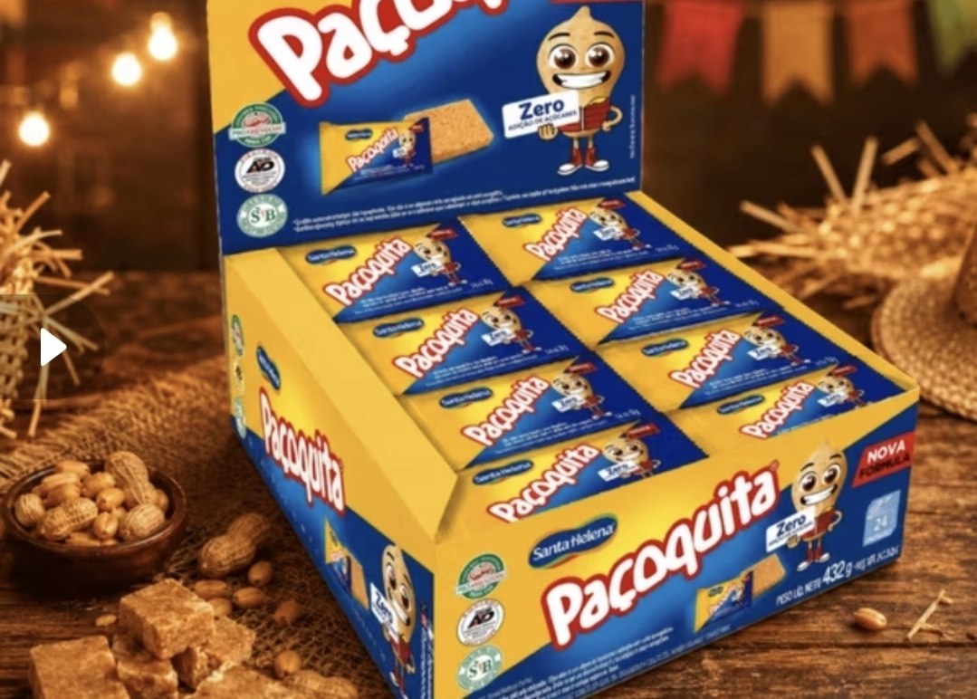 Caixa de papelão amarela da Paçoquita Zero Açúcar Santa Helena aberta, exibindo 24 unidades individuais do doce, com amendoins e paçocas soltas na mesa ao lado.