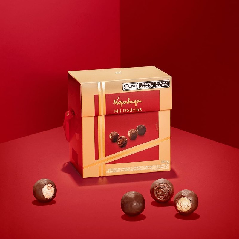 Caixa dourada da Coleção Mil Delícias Kopenhagen de 144g posicionada sobre fundo vermelho, com bombons dispostos ao redor e sobre a embalagem.