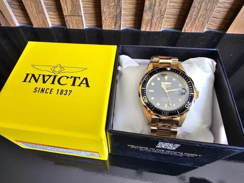 Relógio Invicta masculino com pulseira e caixa douradas e mostrador preto, posicionado dentro de sua caixa amarela original aberta
