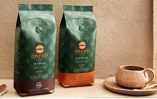 Duas embalagens de Café Orfeu, uma na versão Intenso e outra na versão Classico, ao lado de uma xícara de café em uma mesa.