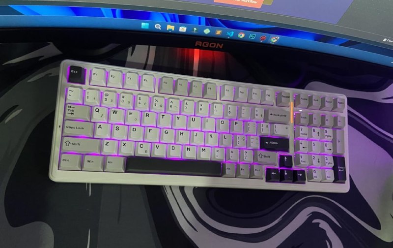 Teclado mecânico Aula F99 Pro branco com teclas cinza, RGB iluminado em roxo, knob laranja ao lado da tecla Backspace, em cima de mousepad sobre mesa gamer