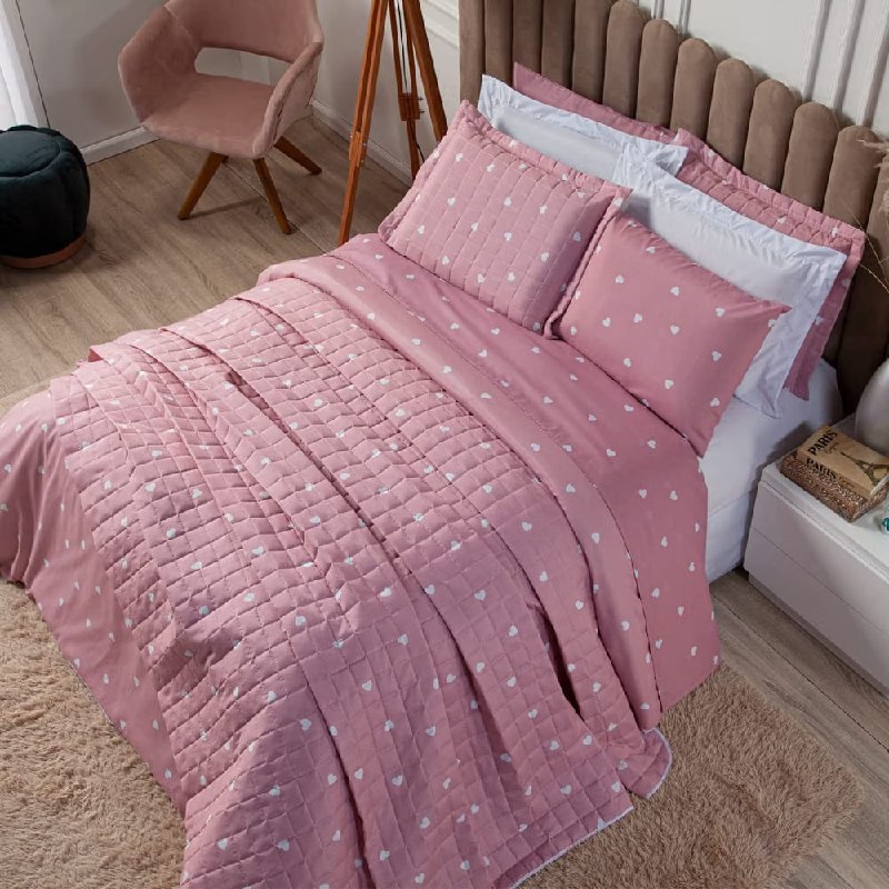 Capa de duvet King da marca Marina Enxovais, modelo Essencialle, na cor rosa com estampa de pequenos corações brancos, sobre uma cama arrumada com travesseiros e almofadas combinando.