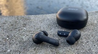 Dois fones de ouvido sem fio pretos da marca QCY modelo MeloBuds HT05 posicionados ao lado de seu estojo de carregamento sobre uma superfície de pedra com água ao fundo.