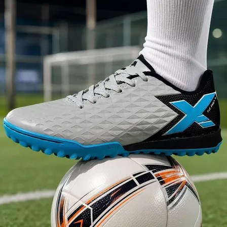 Chuteira Oxn Genio 4 na cor prata com detalhes em azul, posicionada sobre uma bola de futebol em campo de grama sintética.