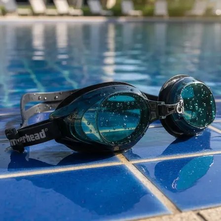 Óculos de natação adulto com lentes escuras e gotas de água, posicionados na borda de uma piscina azul.